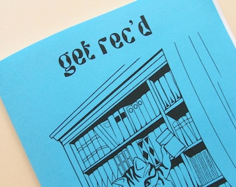 May include: A blue notebook with the words "get rec'd" in black, stylized font. The notebook features a black and white illustration of a cat in front of a bookshelf filled with books.