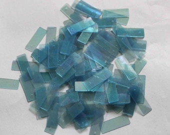 Op de afbeelding: Een stapel kleine, rechthoekige, doorschijnende blauwgroene plastic stukjes. Elk stukje heeft een klein gaatje bij één uiteinde. De stukjes zijn verspreid, waardoor een getextureerd, gelaagd effect ontstaat.