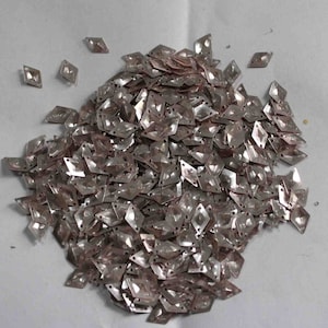 May include: A large pile of silver-colored diamond-shaped sequins. The sequins are scattered on a white surface.