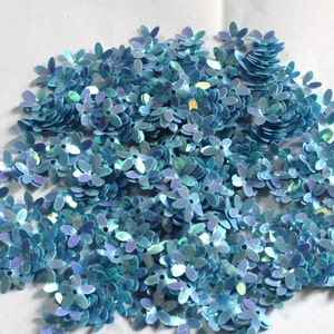 May include: A large pile of light blue iridescent flower-shaped sequins. The sequins are scattered on a white surface.