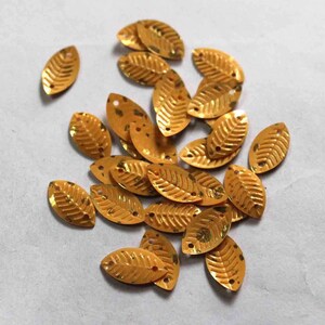 May include: A collection of gold leaf-shaped sequins with a slightly textured surface. The sequins are scattered on a white background.