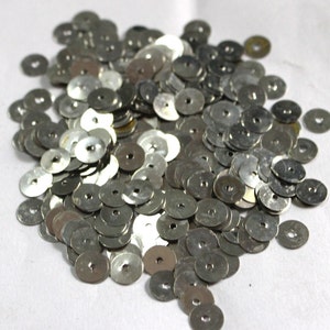 May include: A large pile of silver metal sequins with a hole in the center. The sequins are scattered on a white surface.