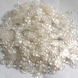 Könnte beinhalten: Ein Haufen durchsichtiger Kunststoffpailletten mit einem blumenartigen Design. Die Pailletten sind klein und haben einen leicht schimmernden Glanz.