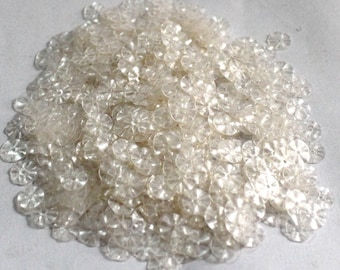 May include: A pile of clear plastic sequins with a flower-like design. The sequins are small and have a slightly iridescent sheen.
