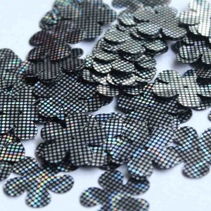 Puede incluir: Una colección de lentejuelas brillantes con forma de flor. Cada lentejuela es negra con un patrón plateado punteado, creando un efecto holográfico. Estos adornos decorativos son ideales para manualidades.