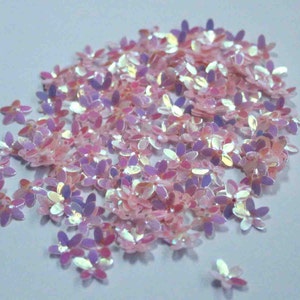Pode incluir: Um grande monte de lantejoulas iridescentes rosa e roxas em forma de flor. As lantejoulas estão espalhadas sobre uma superfície branca.