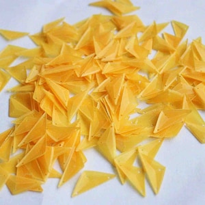 May include: A large pile of yellow triangular sequins. The sequins are scattered on a white surface.