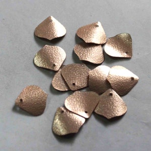 Puede incluir: Un grupo de lentejuelas de color oro rosa en forma de pétalo. Las lentejuelas tienen una superficie texturizada y un pequeño agujero en el centro.