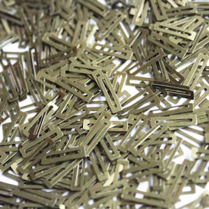 Op de afbeelding: Een grote stapel kleine, goudkleurige metalen stukjes. De stukjes zijn rechthoekig met een kleine opening in het midden. Ze zijn allemaal willekeurig verspreid over een wit oppervlak.