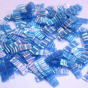 Pode incluir: Um monte de lantejoulas azuis iridescentes com uma textura ondulada e nervurada. As lantejoulas estão espalhadas sobre um fundo azul claro.