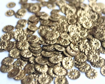 Op de afbeelding: Een stapel gouden metalen versieringen met een decoratieve, gegolfde rand. Elke versiering heeft een klein gaatje in het midden.