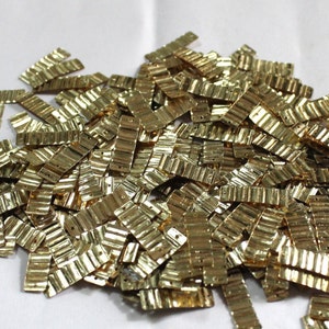 May include: A large pile of gold-colored metal sequins. The sequins are rectangular and have a wavy texture.