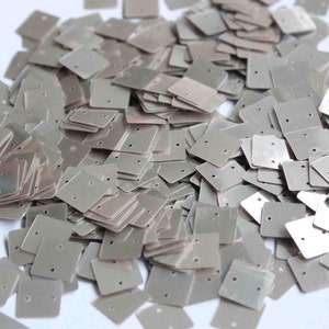May include: A pile of silver square sequins with two holes in each. The sequins are scattered on a white surface.
