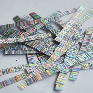 May include: A pile of iridescent, rectangular sequins with a striped pattern. The sequins are silver with a rainbow sheen, and each has a small hole at one end. They are scattered on a light gray surface.