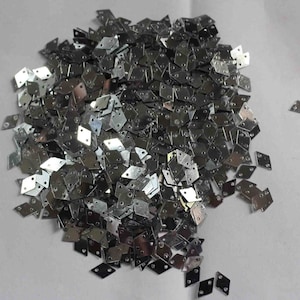 Peut inclure: Un tas de petites pièces métalliques argentées brillantes en forme de losange. Chaque pièce comporte deux petits trous. Les pièces sont dispersées sur une surface blanche.
