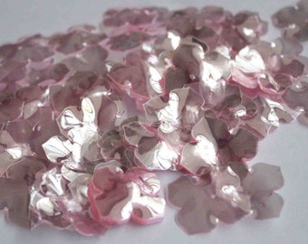 May include: A pile of pink iridescent flower-shaped sequins. The sequins are made of a thin, translucent material and have a slightly shimmery finish.