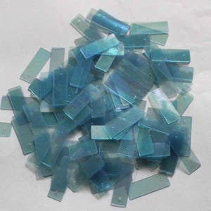 Puede incluir: Un montón de lentejuelas rectangulares iridiscentes de color azul claro. Las lentejuelas están esparcidas sobre una superficie blanca.
