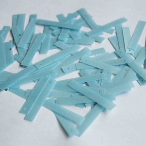 Może przedstawiać: Stosik jasnoniebieskich cekinów w kształcie długich prostokątów. Cękiny są rozrzucone na białym tle.