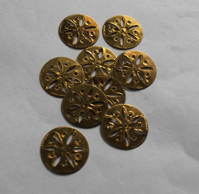 May include: A collection of ten small, round, gold-colored metal embellishments. Each disc features a floral design with cut-out details and a textured edge. The embellishments are scattered on a light gray surface.