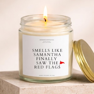 May include: A lit candle in a clear glass jar with a gold lid. The label reads "Smells Like Samantha Finally Saw The Red Flags" with a red flag graphic. The candle is hand-poured in the USA.