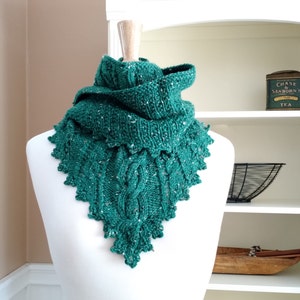 Puede incluir: Bufanda verde tejida a mano con un diseño de punto de cable texturizado y un borde festoneado. La bufanda se muestra en un maniquí blanco y tiene motas blancas en todo el hilo. La bufanda tiene forma triangular.
