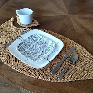 May include: A tan knitted placemat with a white plate and a smaller patterned plate on top. A silver fork, knife, and spoon are placed on the placemat.