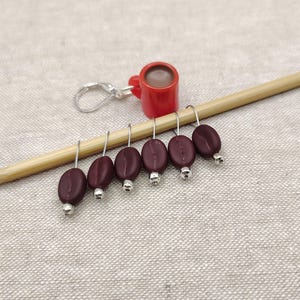 May include: A set of six stitch markers featuring dark brown coffee bean charms with silver-toned beads and a single red coffee mug charm, all suspended from silver-toned metal loops and resting on a light-colored wooden knitting needle.