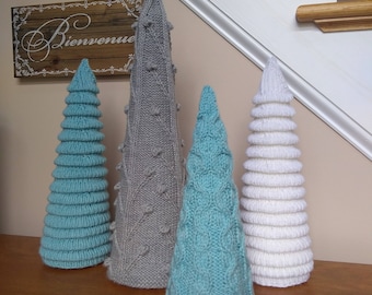 Puede incluir: Cuatro árboles de Navidad tejidos en diferentes colores y patrones. Los árboles son grises, azul verdoso y blanco. Los árboles están de pie sobre una superficie de madera. El letrero detrás de los árboles dice "Bienvenue".