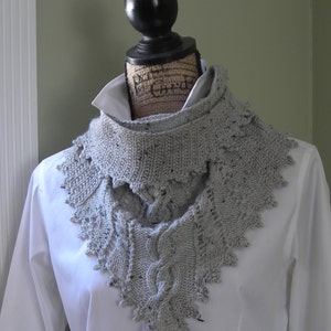 Puede incluir: Una bufanda de punto gris con un patrón de trenza y un borde de encaje. La bufanda se muestra sobre una camisa blanca.