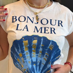 Puede incluir: Una camiseta blanca de manga corta con el texto azul oscuro BONJOUR LA MER sobre un gráfico grande de una concha marina en azul y dorado. La prenda se combina con un collar de varias capas que incluye una cadena dorada y cuentas blancas con un pez.