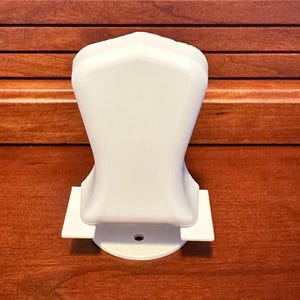 May include: A white, plastic, decorative object with a rounded top and a flat base. The object has a unique shape and is set against a brown wooden background. The base has a small hole in the center.