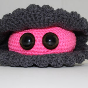 Puede incluir: Una almeja de crochet rosa con grandes ojos negros. La almeja está cerrada y tiene una concha de crochet gris.