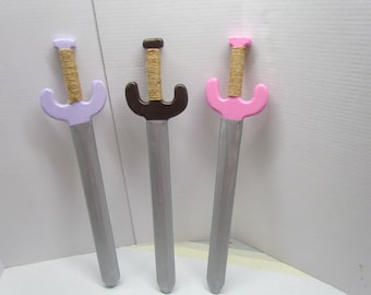May include: Three wooden toy swords with different colored handles. The swords have a silver blade and a brown wrapped handle. The handles are lavender, brown, and pink.