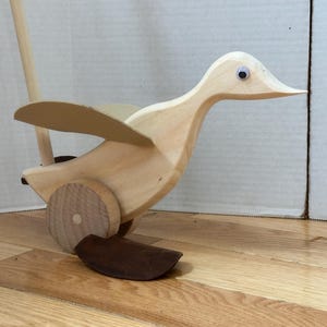 Puede incluir: Un juguete de madera con forma de pato para tirar, con un mango largo, alas y ruedas. El pato es de madera clara con detalles marrones en las patas y las ruedas. El pato tiene ojos saltones y está diseñado para niños pequeños.