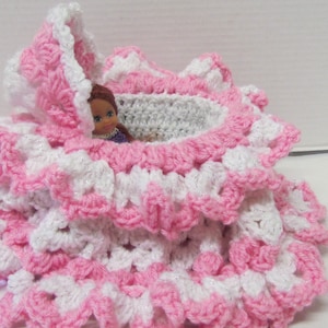 May include: A pink and white crocheted doll crib with a doll inside. The crib is made of multiple layers of crocheted ruffles.