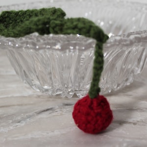 Pode incluir: Uma cereja de crochê feita à mão, com um caule verde e uma fruta vermelha. A cereja está posicionada em frente a uma tigela de vidro transparente com um design texturizado. O fundo é uma superfície semelhante a mármore.