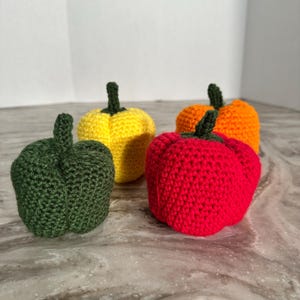 May include: Four crocheted bell peppers in different colors: green, yellow, orange, and red. The peppers are arranged in a row on a light gray surface.