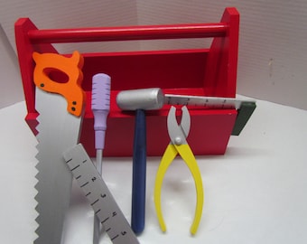 May include: A red wooden toy toolbox with a handle, containing a saw, a screwdriver, a hammer, pliers, and a ruler. The tools are made of wood and painted in various colors.