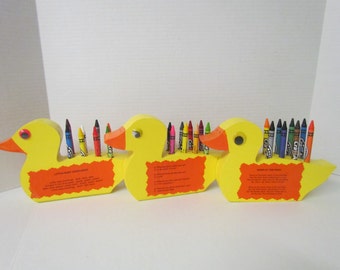 May include: Three yellow duck-shaped crayon holders with orange beaks and eyes. Each duck holds a collection of colorful crayons in its back. The ducks have orange labels with text. A fun and functional desk accessory for kids.