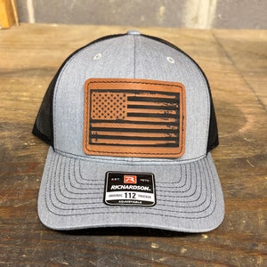 May include: A gray and black trucker hat with a brown leather patch featuring a distressed American flag design. The patch also includes the text "EST. 1970 RICHARDSON. ORIGINAL TRUCKER 112 ADJUSTABLE".