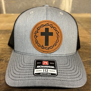 May include: A gray and black trucker hat with a brown leather patch featuring a cross inside a thorned circle. The patch also has the text "Richardson" and "112" on it.