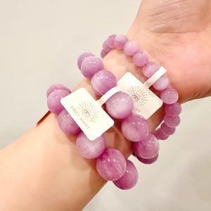 May include: Three bracelets with large, round, light purple beads are displayed on a wrist. Each bracelet has a tag that reads "FIRST SIGHT" with an eye symbol. The beads have a polished, glossy finish.