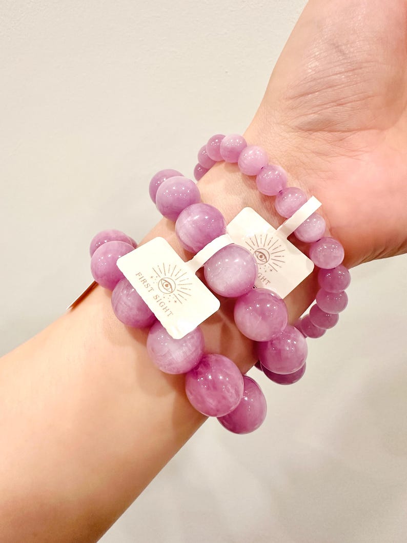 May include: Three bracelets with large, round, light purple beads are displayed on a wrist. Each bracelet has a tag that reads "FIRST SIGHT" with an eye symbol. The beads have a polished, glossy finish.