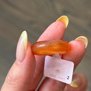 May include: A smooth, translucent orange resin ring with subtle swirling patterns throughout the material. Attached to the band is a small white rectangular tag featuring a graphic of an eye with sun rays and the text FIRST SIGHT followed by the number 2.