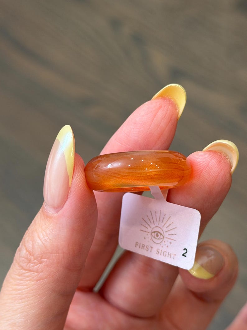 May include: A smooth, translucent orange resin ring with subtle swirling patterns throughout the material. Attached to the band is a small white rectangular tag featuring a graphic of an eye with sun rays and the text FIRST SIGHT followed by the number 2.