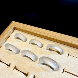 Könnte beinhalten: Sechs durchscheinende, hellgraue Ringe, die in einer Holzschale mit beigem Stofffutter ausgestellt sind. Die Ringe haben ein glattes, abgerundetes Design und sind gleichmäßig in den Fächern der Schale angeordnet. Der Hintergrund ist dunkel.