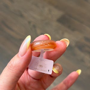 May include: A smooth, translucent orange ring with subtle white swirling patterns and tiny embedded glitter accents. A small white rectangular tag is attached to the band, featuring a graphic of an eye with rays and the text FIRST SIGHT 1.