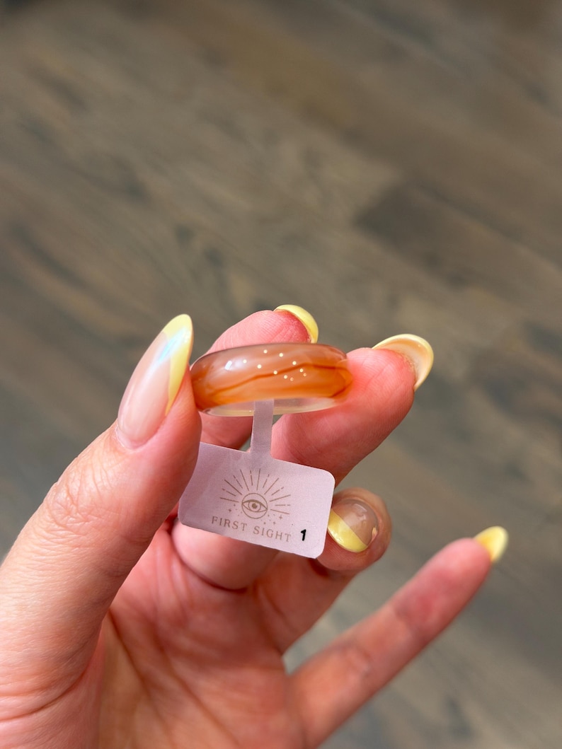 May include: A smooth, translucent orange ring with subtle white swirling patterns and tiny embedded glitter accents. A small white rectangular tag is attached to the band, featuring a graphic of an eye with rays and the text FIRST SIGHT 1.