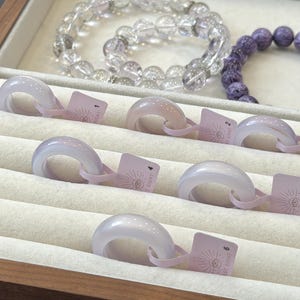 May include: Six smooth, pale lavender-grey stone rings arranged in a display tray, each featuring a small light pink tag numbered one through six. In the background, two beaded bracelets are visible, one clear and one deep purple.