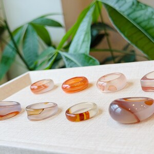 May include: A collection of eight smooth, polished rings crafted from translucent banded agate stone. Each ring features unique natural color variations, including shades of orange, white, brown, and grey, with distinct swirling patterns and polished surfaces.